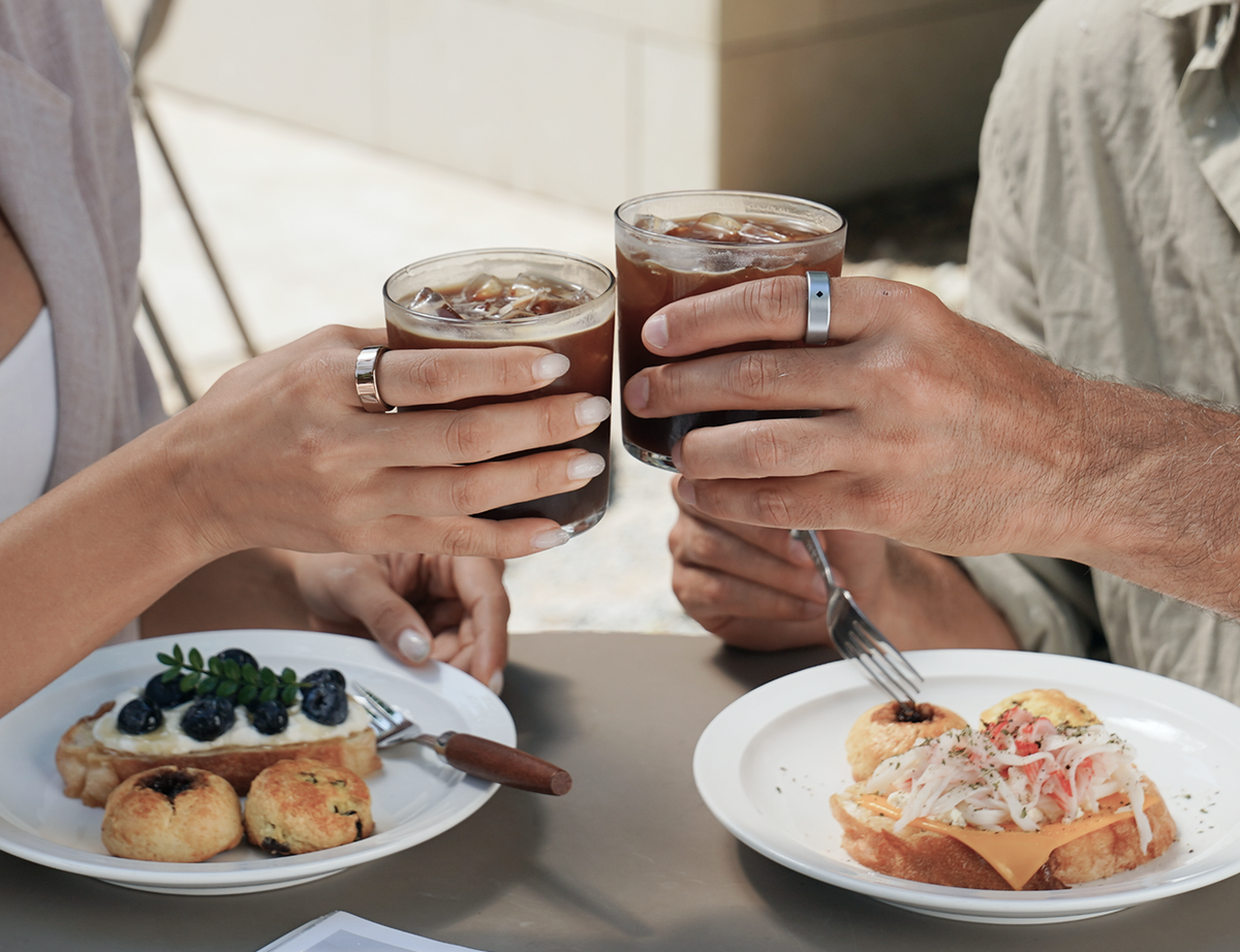 Two people wearing AIVELA rings sharing iced coffee at a sunlit table
